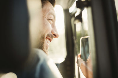 Happy man looking away while photographing through smart phone in car
