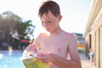 Shirtless boy opening packet against swimming pool in sunny day