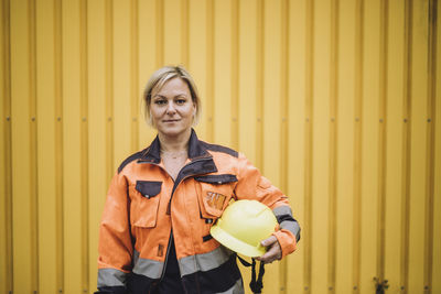 Portrait of mature female construction worker with hardhat against yellow metal wall