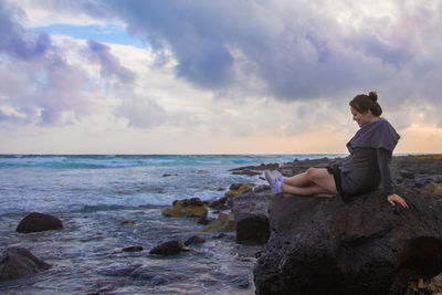 Woman sitting on rock at beach against cloudy sky during sunset