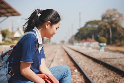 Side view of young woman sitting on railroad track against sky