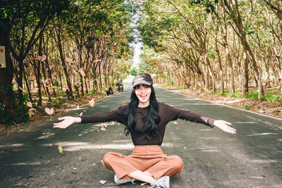 Portrait of smiling young woman throwing leaves while sitting on road amidst trees in forest