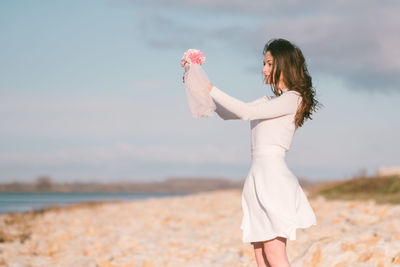 Portrait of young woman standing at beach against sky