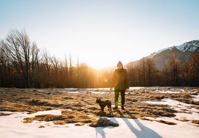 Dog owner and french bulldog on snow covered landscape against mountain and sky at sunset