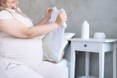 Happy pregnant woman with big belly holding baby clothes in hands at home