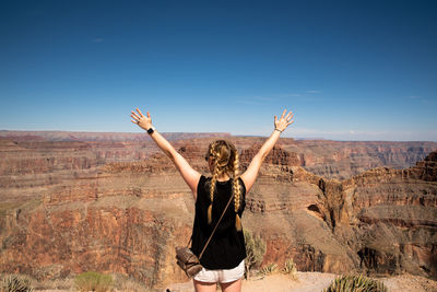 Rear view of woman with arms raised standing against rock formation and sky
