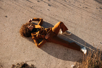 From above of curly haired woman in trendy outfit lying on asphalt road in evening sunlight in countryside