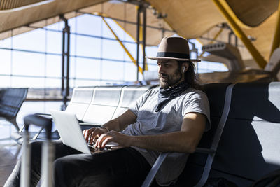 Side view of focused man with long hair in stylish outfit with hat sitting in a bench next to glass wall working on a laptop while waiting for flight in airport lounge