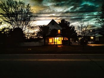 Illuminated street by building against sky at dusk