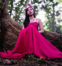 Low angle view of woman in red evening gown sitting by tree trunk