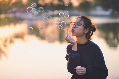 Young woman blowing bubbles while standing against lake during sunset