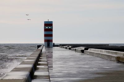 Wet pier leading towards lighthouse by sea against sky