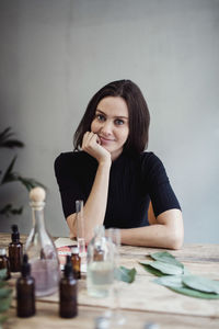 Portrait of confident female owner sitting with hand on chin at table against wall in perfume workshop
