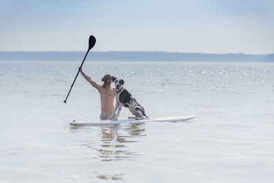 Shirtless man with dog paddleboarding in sea against sky