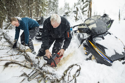 Elderly man and son using chainsaw to clear trails while snowmobiling.