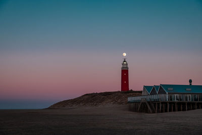 Lighthouse amidst sea and buildings against sky during sunset