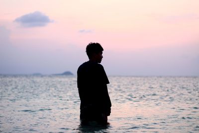 Rear view of man looking away while standing in sea against sky during sunset