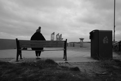 Rear view of woman sitting on bench at footpath against cloudy sky