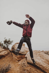 Low angle view of man holding camera while jumping on rocks against sky