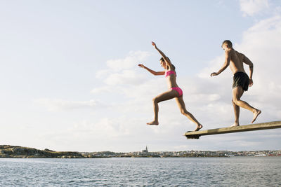 Full length of young couple diving into lake against cloudy sky