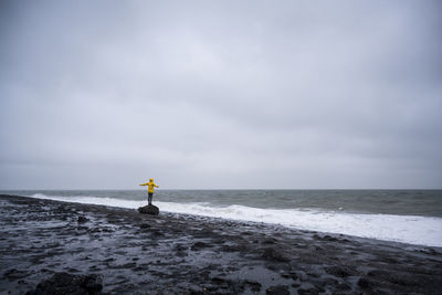 Woman with arms outstretched standing on rock at sea shore against cloudy sky