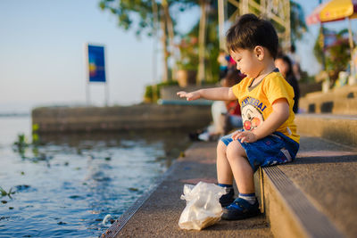 Side view of cute baby boy throwing food in lake while sitting on steps during sunset