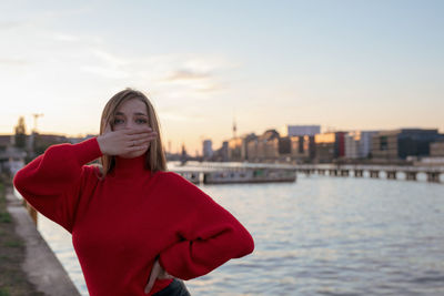 Portrait of young woman with hands covering mouth standing by river in city against sky