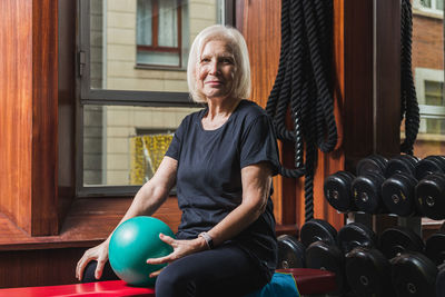 Happy senior female athlete with smart watch sitting on bench with small exercise ball against dumbbells in gymnasium looking at camera