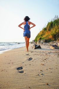 Rear view of woman with hands on hip walking on shore at beach against sky