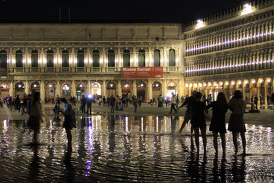 People enjoying water fountain against illuminated buildings