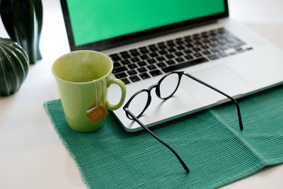 Laptop on table with green screen and hot cup of tea desk with accessories and devices for home work