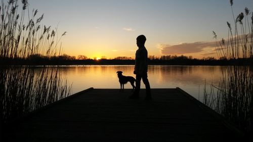 Silhouette boy with dog on pier over lake against sky during sunset