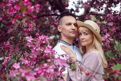 Portrait man and a woman in hat stand by a blooming pink cherry tree in summer