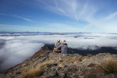 Rear view of man and woman sitting on rock against sky