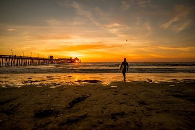 Rear view of surfer walking at beach against sky during sunset