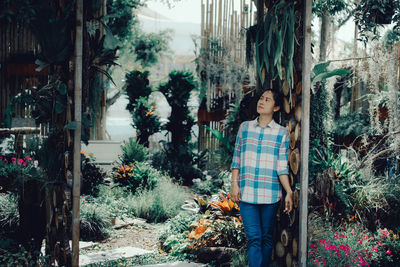 Woman looking away while standing by wall against plants in greenhouse