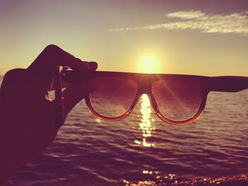 Close-up of hand holding sunglasses against sky during sunset