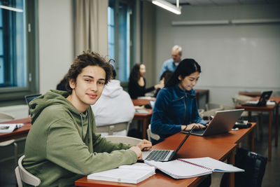 Portrait of smiling teenage boy using laptop at desk with classmates studying in classroom