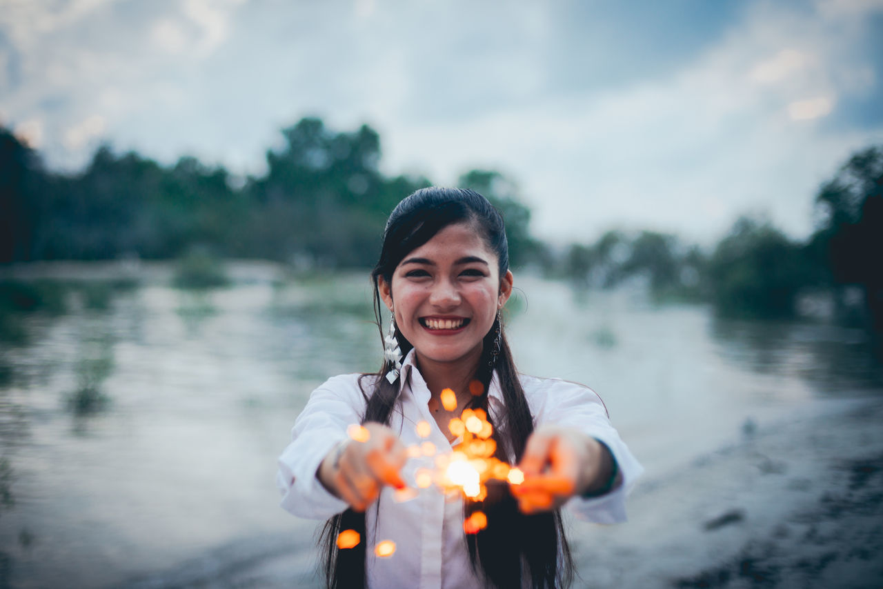 PORTRAIT OF HAPPY YOUNG WOMAN STANDING AGAINST LAKE