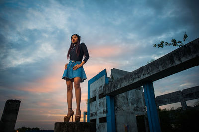 Low angle view of young woman against abandoned building during sunset