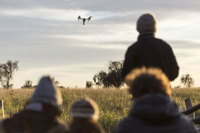 Rear view of people on field with drone flying against cloudy sky