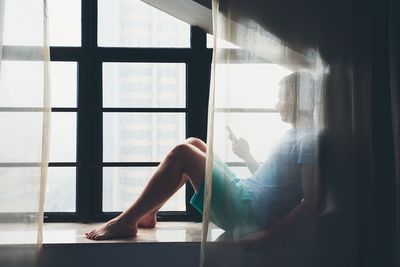 Full length of man behind curtain using phone on window sill at home