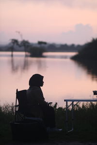 Rear view of silhouette man sitting by lake against sky during sunset