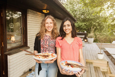 Portrait of happy teenage girls holding salad bowls outside house at yard