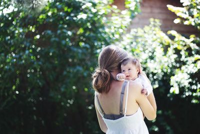 Rear view of mother carrying toddler daughter against plants