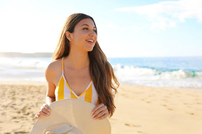 Smiling young woman holding hat while standing at beach