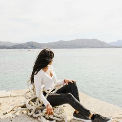 Woman sitting on mountain by lake against sky