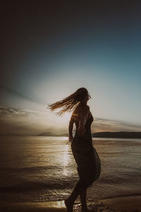 Rear view of woman standing at beach against sky during sunset