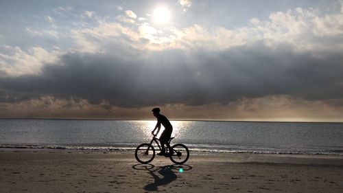 Man riding bicycle at beach against cloudy sky