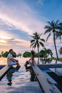 People sitting by swimming pool against sky during sunset